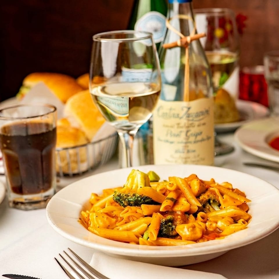 A table set with Italian dishes, including penne pasta, lasagna, and bread, with wine and drinks.