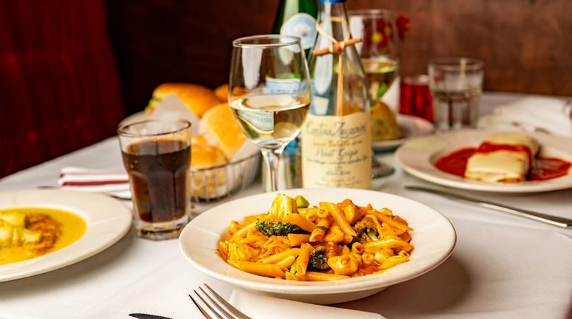 A table set with Italian dishes, including penne pasta, lasagna, and bread, with wine and drinks.