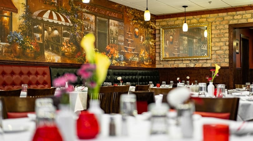 Restaurant dining room with a large mural, brick wall, and set tables with red accents.