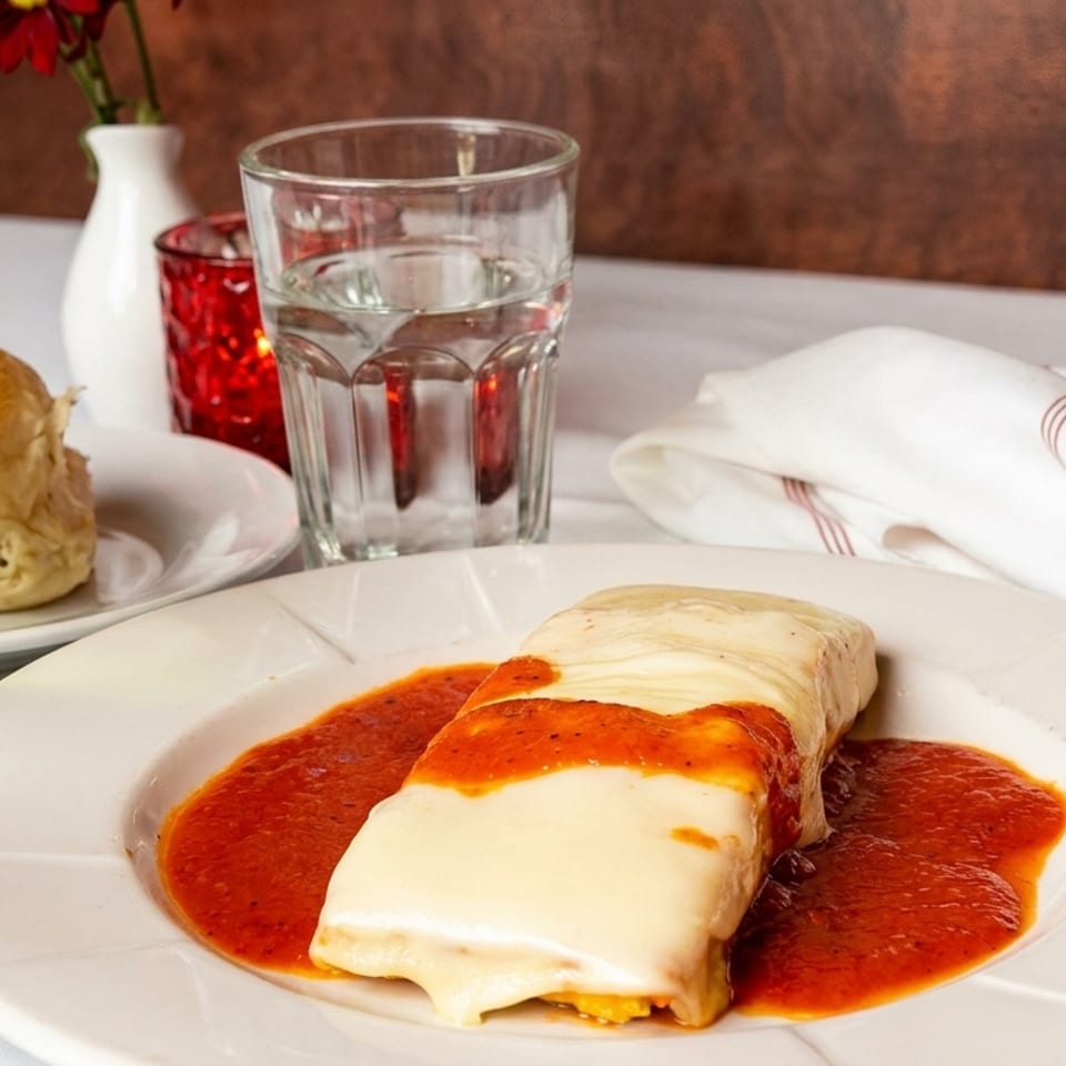 A cheese-covered lasagna with red sauce, a bread roll, white wine, and water on a white tablecloth.