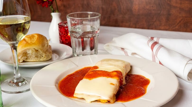 A cheese-covered lasagna with red sauce, a bread roll, white wine, and water on a white tablecloth.