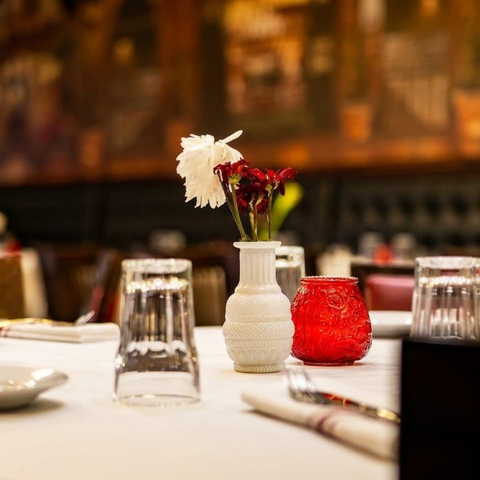 A restaurant table set with white plates, glasses, silverware, and a vase of flowers.