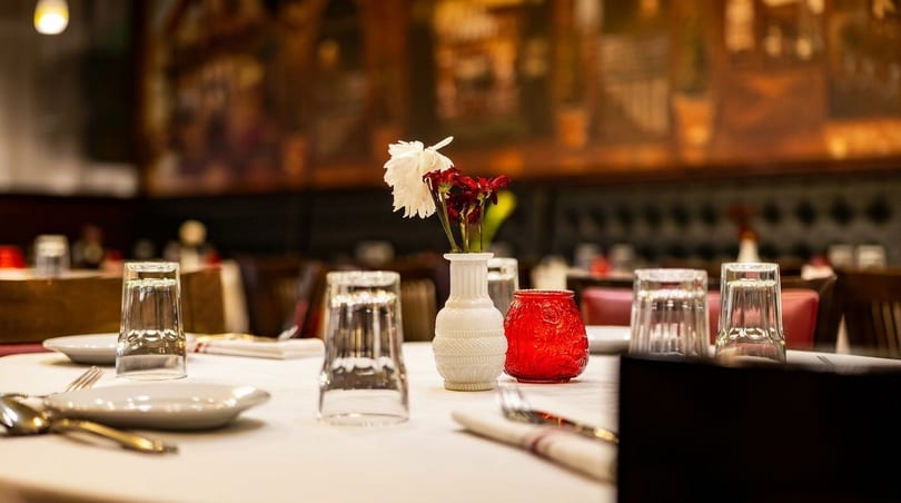 A restaurant table set with white plates, glasses, silverware, and a vase of flowers.