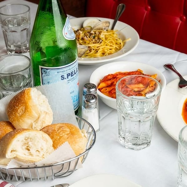 Table set with Italian pasta dishes, bread rolls, water glasses, and a green bottle of sparkling water.