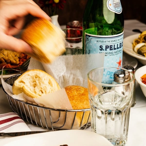 A hand reaching for bread from a basket on a table with Italian pasta dishes and sparkling water.