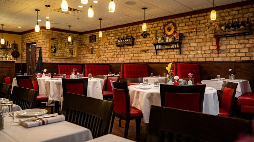 An empty Italian restaurant dining room with brick walls, red booths, and white-clothed tables.