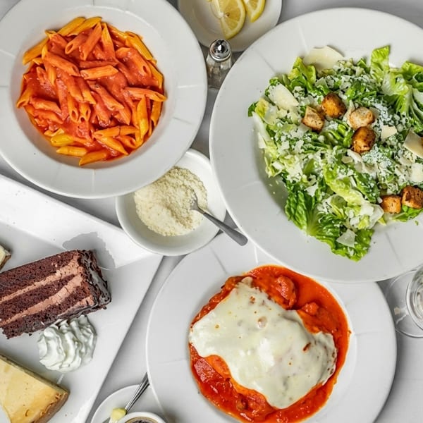 Overhead view of Italian dishes: penne pasta, chicken parmesan, Caesar salad, and dessert slices.